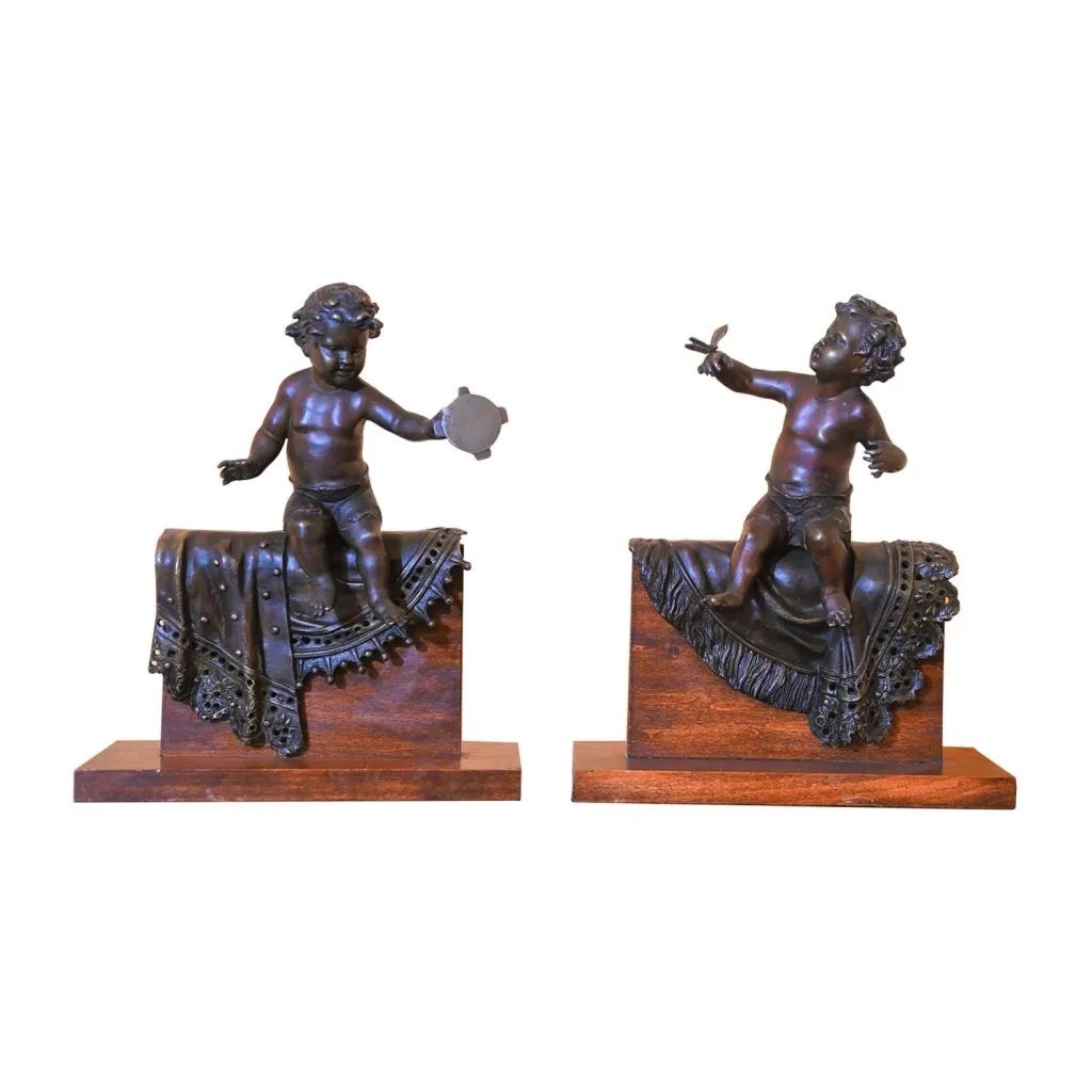 Pair Of Bronze Sculptures With A Burnished  Patina. Two Cherubs Lying On A Richly Decorate Bla 